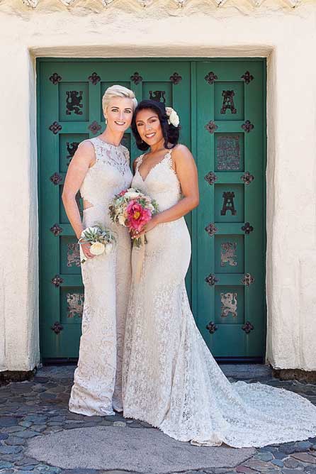 two lesbian brides at rustic Mexican wedding in a lace BHLDN wedding gown and lace jumpsuit with exposed back and holding bouquets standing in a garden
