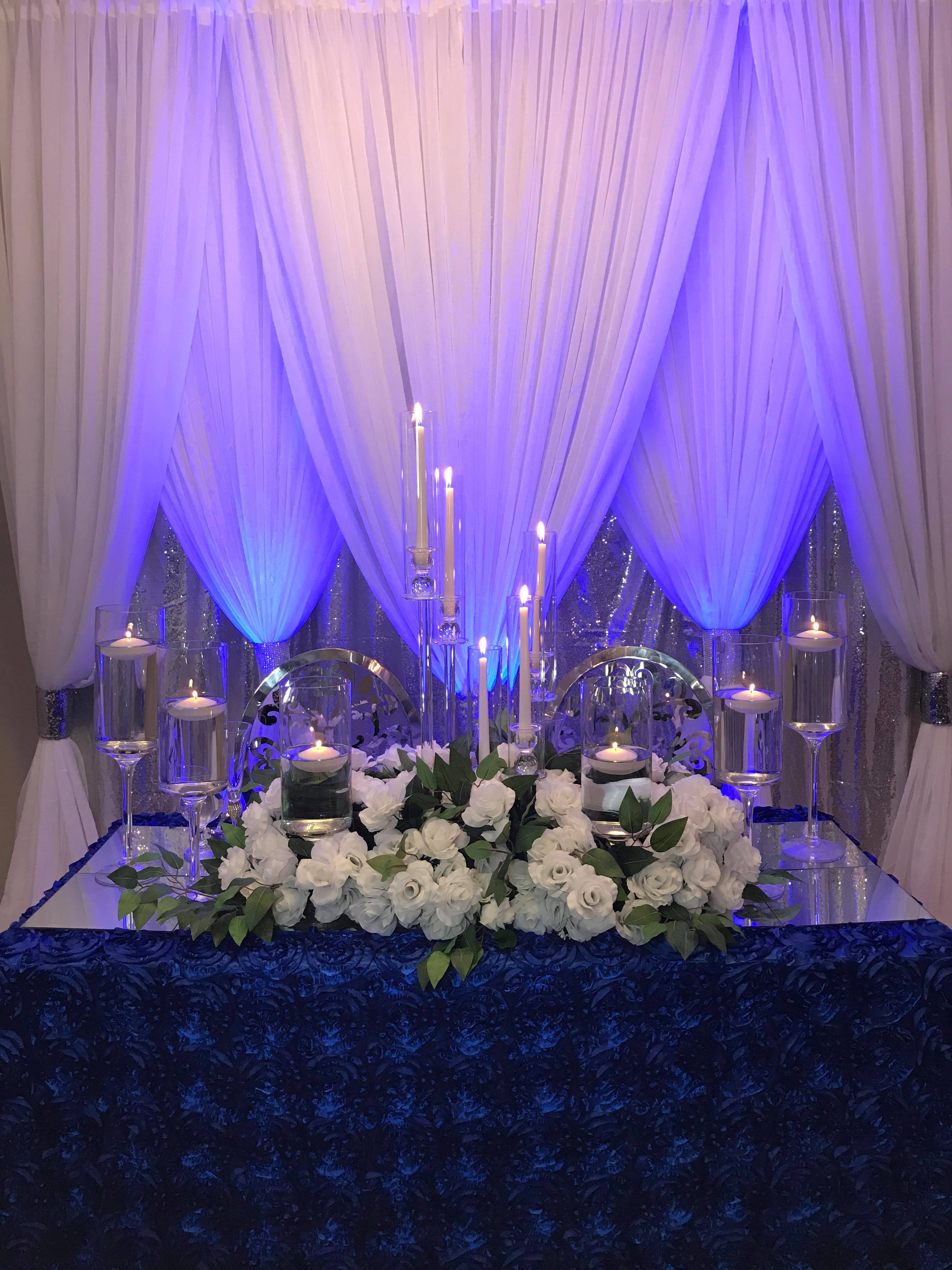 A sweetheart table with white florals and candles, set against a backdrop of white drapes. ADTR.