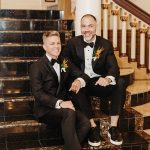 two grooms sit on stairs, smiling at the camera