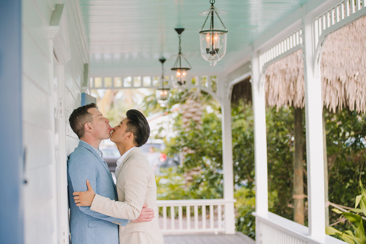 Intimate Florida Keys tropical wedding two grooms blue tan tuxedo sitting cheers wine glasses kisses porch