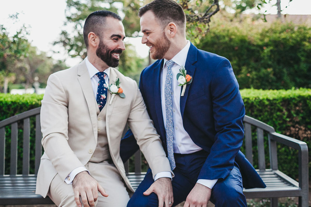 Historic hometown wedding in Pasadena, California two grooms blue tux gray tux boutonnieres holding hands bench