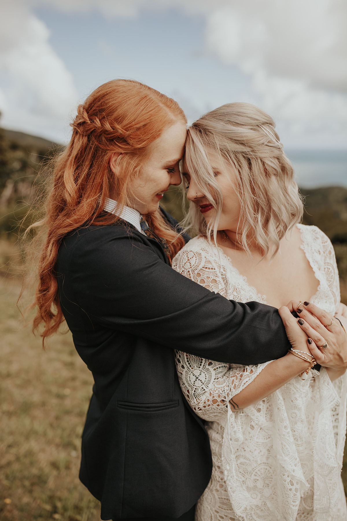 Texas couple elopes to LGBTQ+ friendly New Zealand and their warm, moody beach wedding photos are to die for two brides white lace dress red hair black suit bow tie