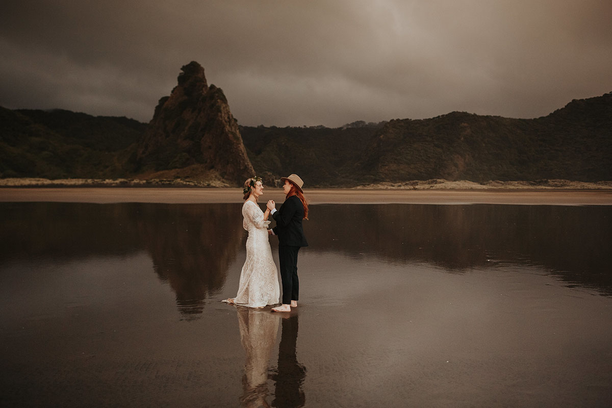 Texas couple elopes to LGBTQ+ friendly New Zealand and their warm, moody beach wedding photos are to die for two brides white lace dress red hair black suit bow tie