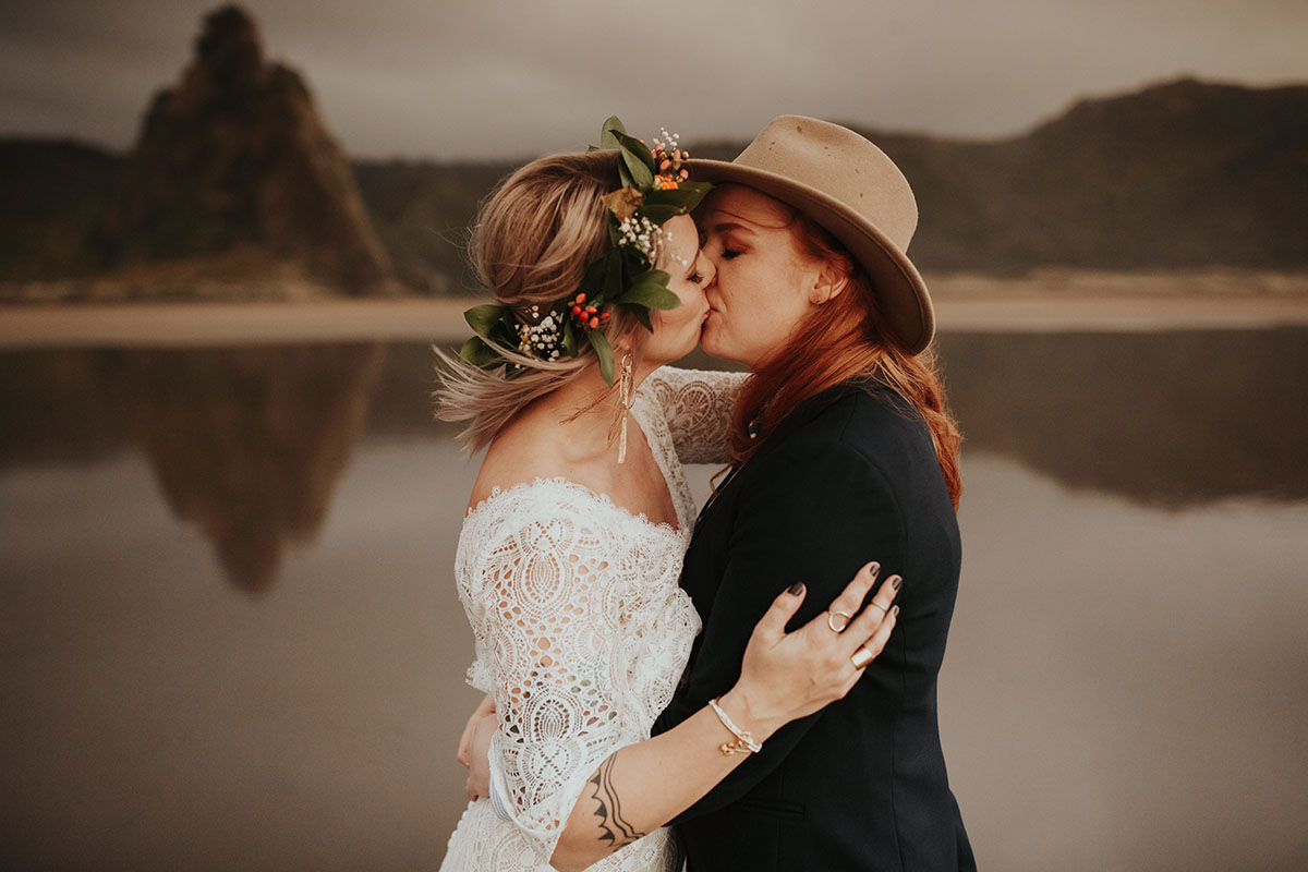 Texas couple elopes to LGBTQ+ friendly New Zealand and their warm, moody beach wedding photos are to die for two brides white lace dress red hair black suit bow tie kiss