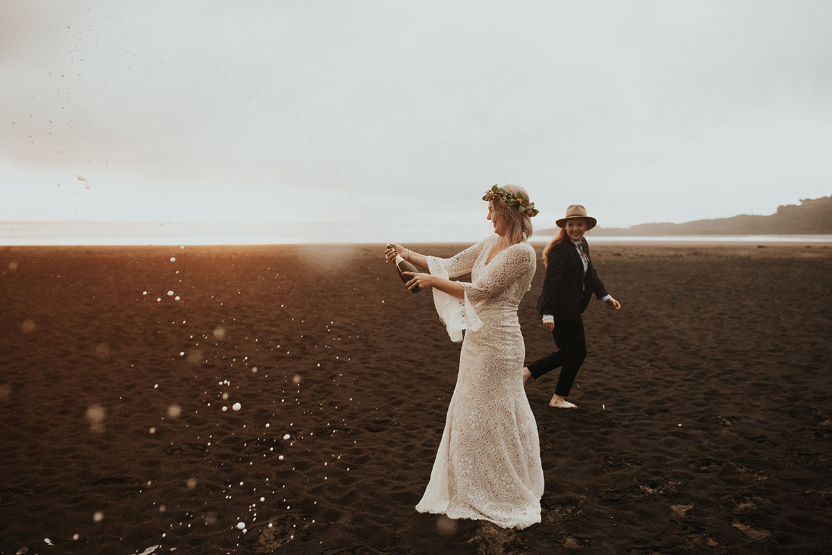 Texas couple elopes to LGBTQ+ friendly New Zealand and their warm, moody beach wedding photos are to die for two brides white lace dress red hair black suit bow tie champagne