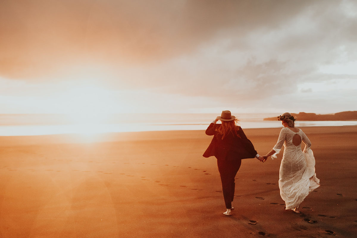 Texas couple elopes to LGBTQ+ friendly New Zealand and their warm, moody beach wedding photos are to die for two brides white lace dress red hair black suit bow tie sunset