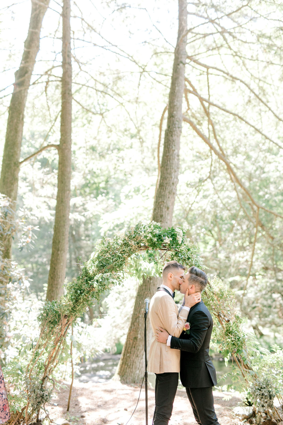 Intimate summer wedding in the woods surrounded by nature two grooms black tux tan tux matching bow ties kiss