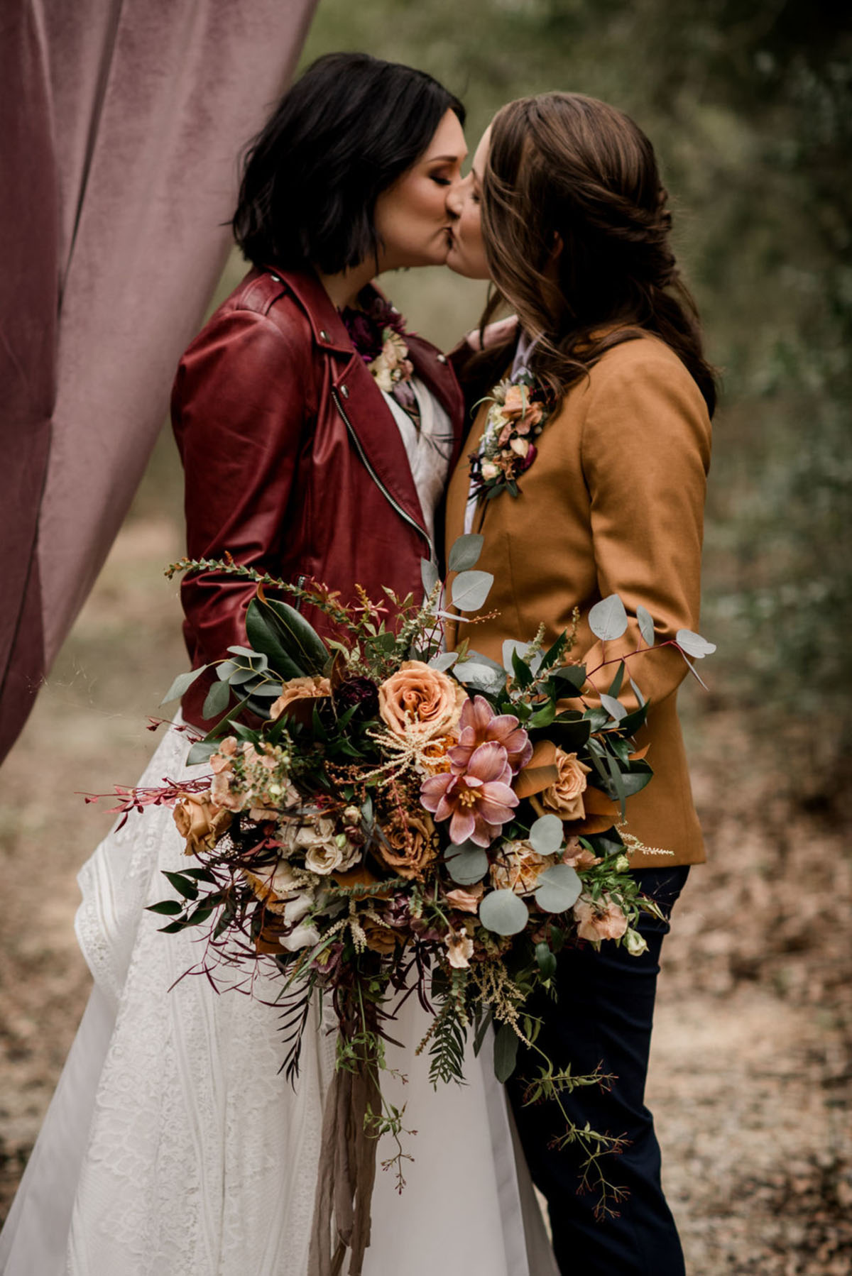 Floral bohemian forest wedding inspiration on a bridge two brides lesbian wedding long white dress tan suit jacket red leather jacket romantic moody kiss