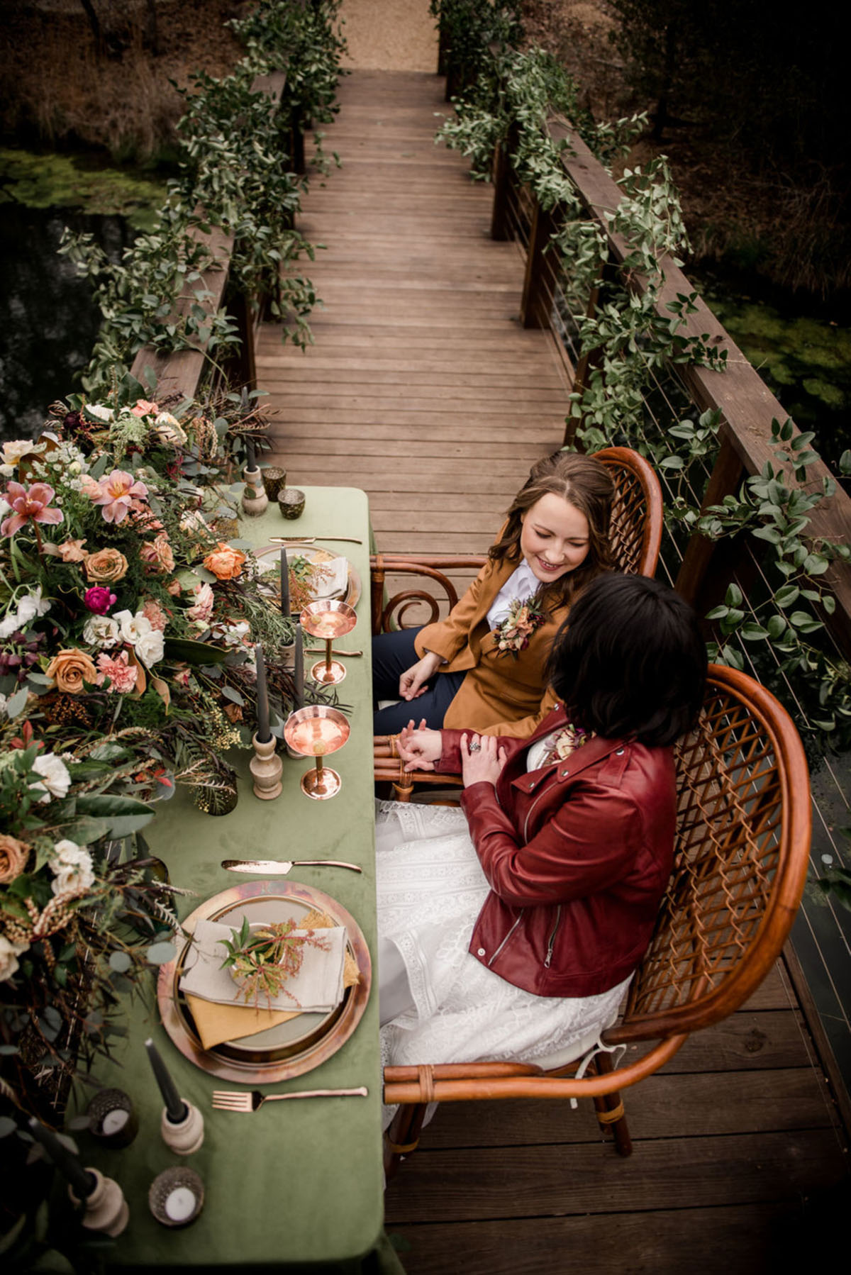Floral bohemian forest wedding inspiration on a bridge two brides lesbian wedding long white dress tan suit jacket red leather jacket romantic moody sweetheart table