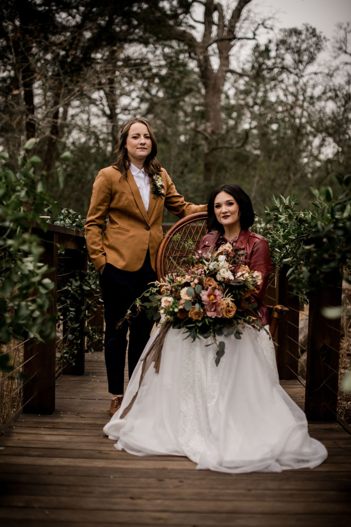 Floral bohemian forest wedding inspiration on a bridge two brides lesbian wedding long white dress tan suit jacket red leather jacket romantic moody