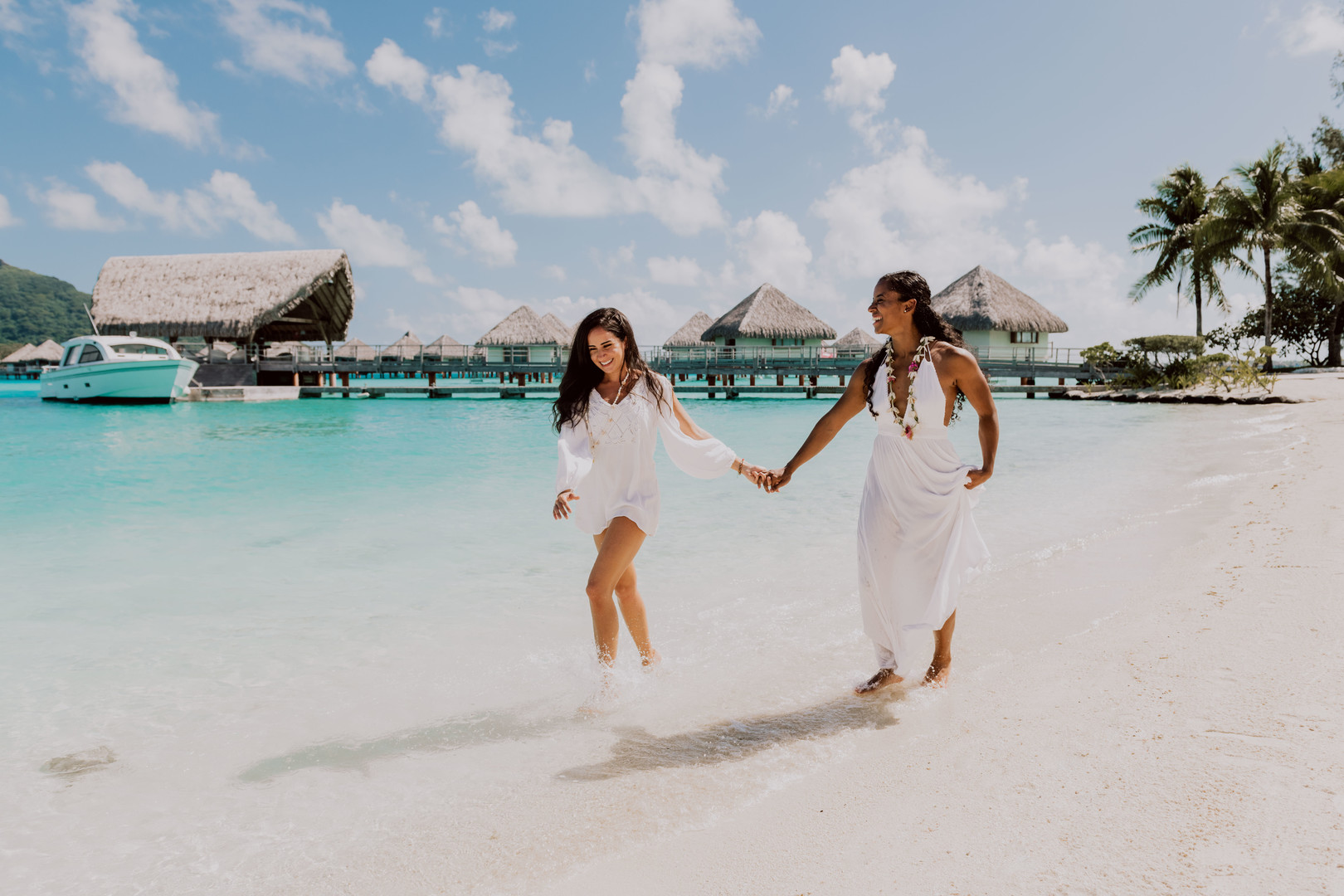 Beach honeymoon photos in Bora Bora two brides white dresses clear blue oceans palm trees holding hands
