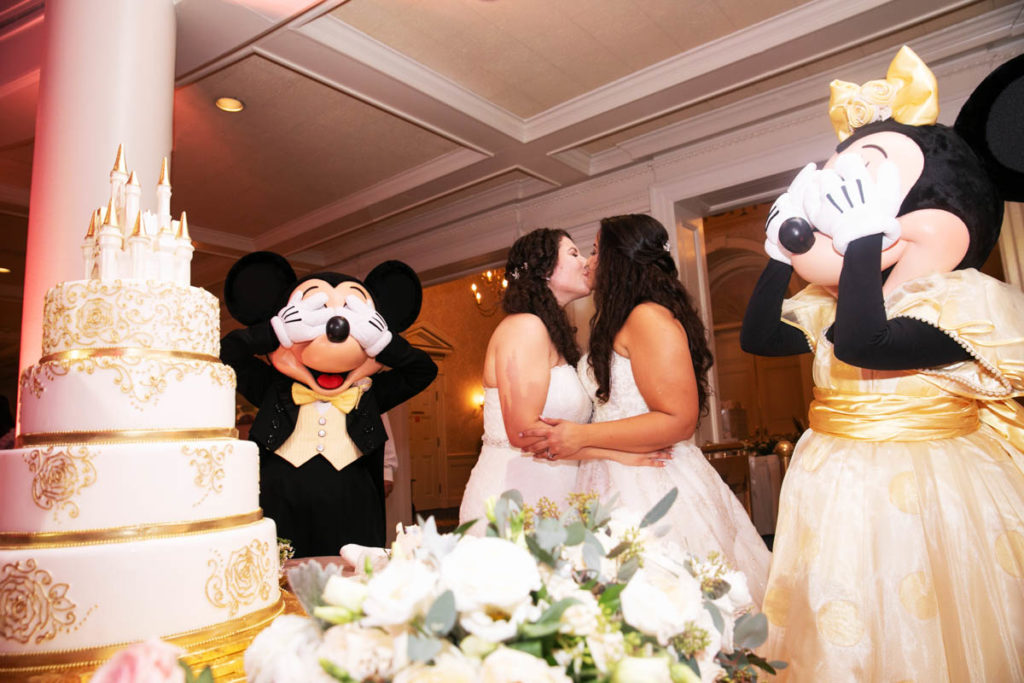 two brides cut wedding cake while Mickey Mouse and Minnie Mouse pretend to hide their eyes