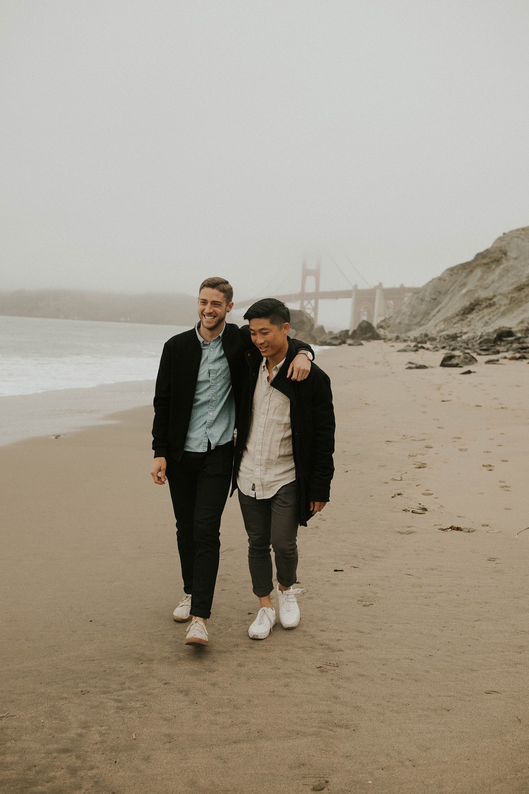 Fun, moody beach couple photos in San Francisco, California LGBTQ+ couples portraits two grooms Baker Beach Golden Gate Bridge
