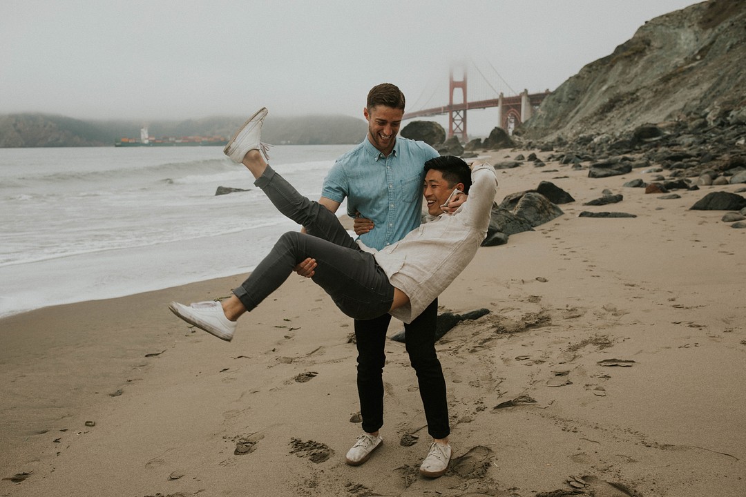 Fun, moody beach couple photos in San Francisco, California LGBTQ+ couples portraits two grooms Baker Beach Golden Gate Bridge