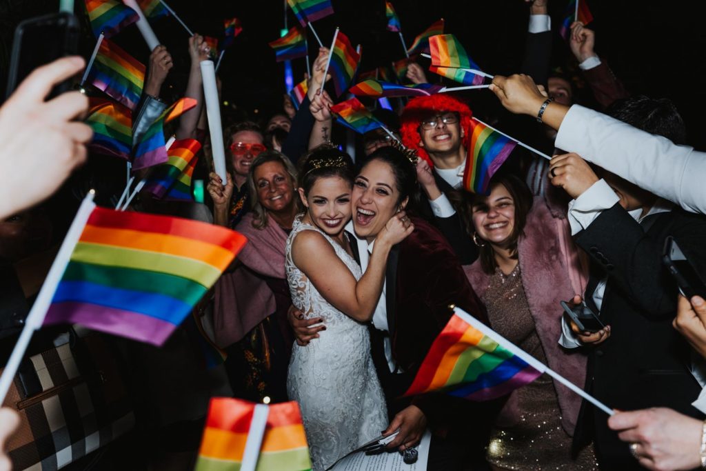 lesbian couple surrounded by family with Pride flags at LGBTQ+ wedding
