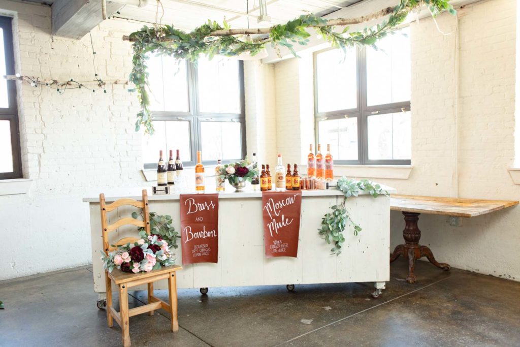 An all-female vendor team created this gorgeous burgundy and gold LGBTQ+ wedding inspiration at The White Room in Worcester, Massachusetts. The venue is situated in a historic mill building. The models are a real-life couple who are getting married this October. We love the burgundy contrasted with blush, rusty orange and the leather and gold touches.