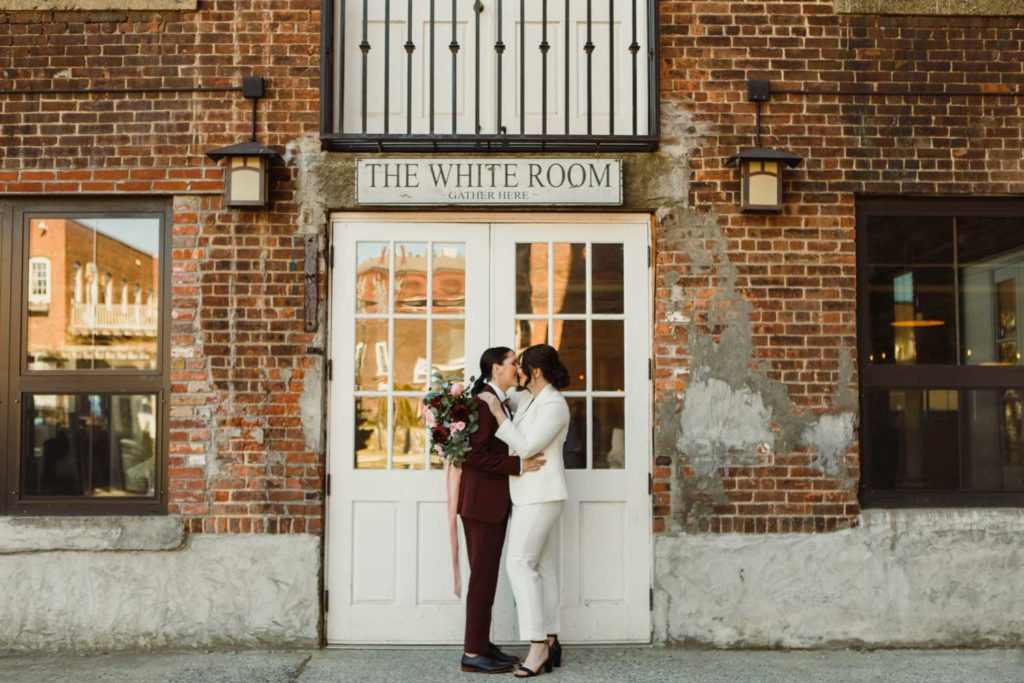 A n all-female vendor team created this gorgeous burgundy and gold LGBTQ+ wedding inspiration at The White Room in Worcester, Massachusetts. The venue is situated in a historic mill building. The models are a real-life couple who are getting married this October. We love the burgundy contrasted with blush, rusty orange and the leather and gold touches.