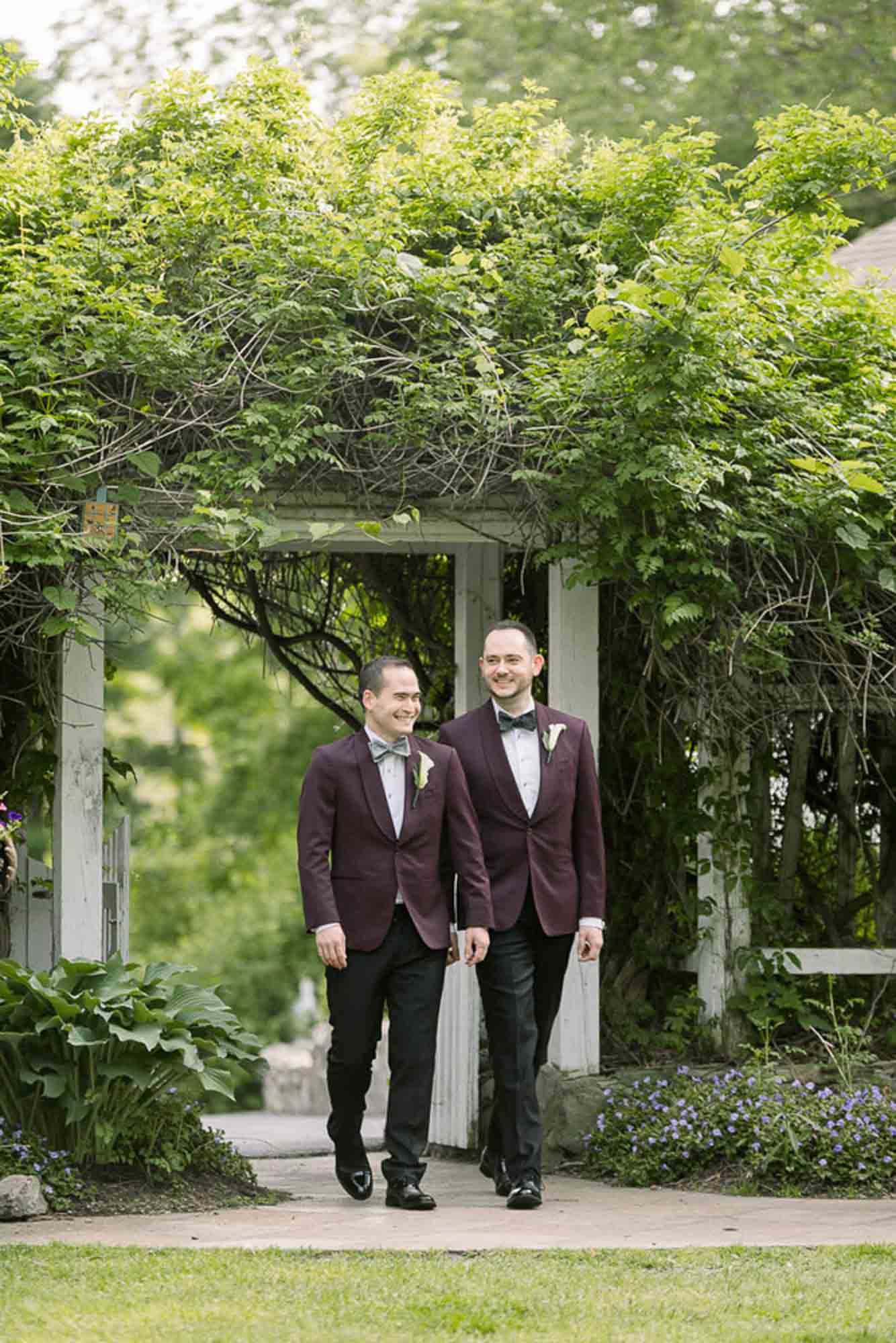 grooms walking into wedding together hand in hand wearing eggplant purple suit jackets LGBTQ+ wedding featured on Equally Wed magazine with two grooms Brian + Brett: Hudson Valley garden wedding with shades of purple Upasana Mainali Photography outdoor wedding ceremony