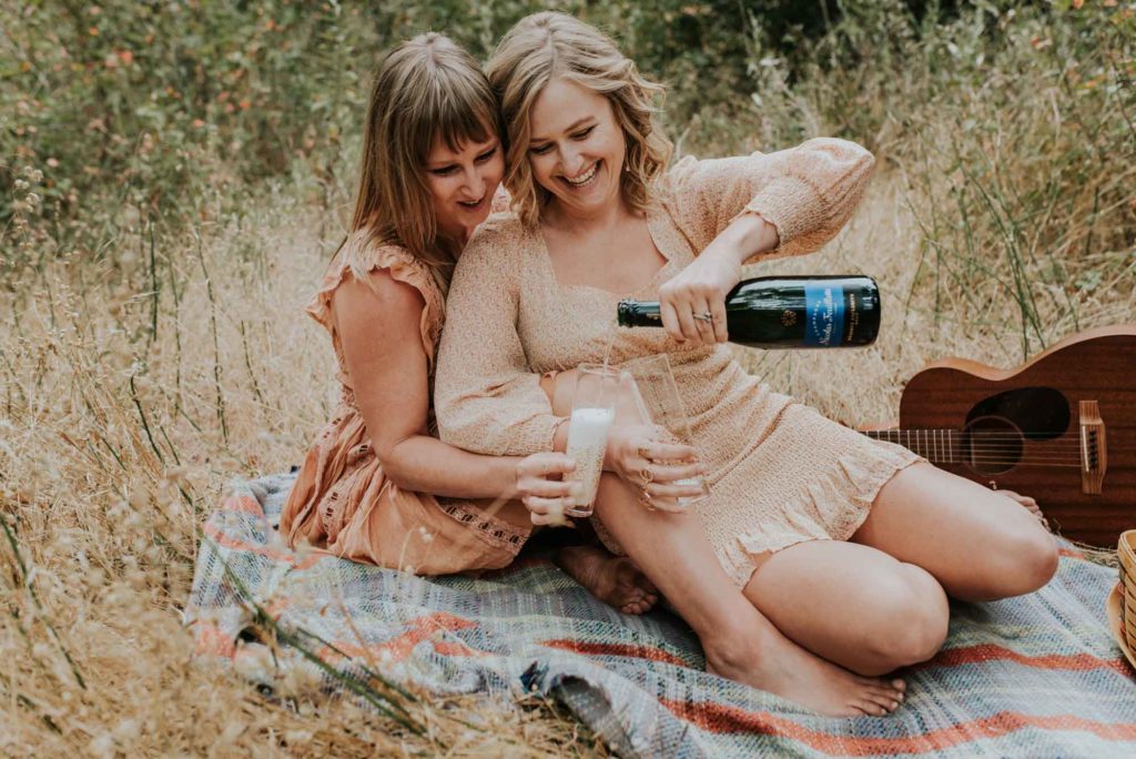 Magical engagement photo session in Big Sur. Photography by Heather K. Purdy. Featured on Equally Wed, the leading LGBTQ+ wedding magazine and wedding vendor directory of LGBTQ+ inclusive wedding pros. pictured: Two women in love having a picnic on the coast of California with a champagne toast and a guitar in the background