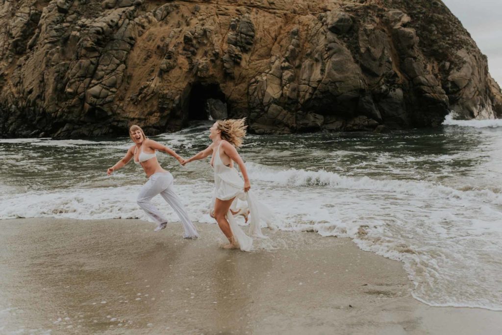 Magical engagement photo session in Big Sur. Photography by Heather K. Purdy. Featured on Equally Wed, the leading LGBTQ+ wedding magazine and wedding vendor directory of LGBTQ+ inclusive wedding pros. romantic sexy image includes two blonde women in love running hand in hand in white outfits on beach