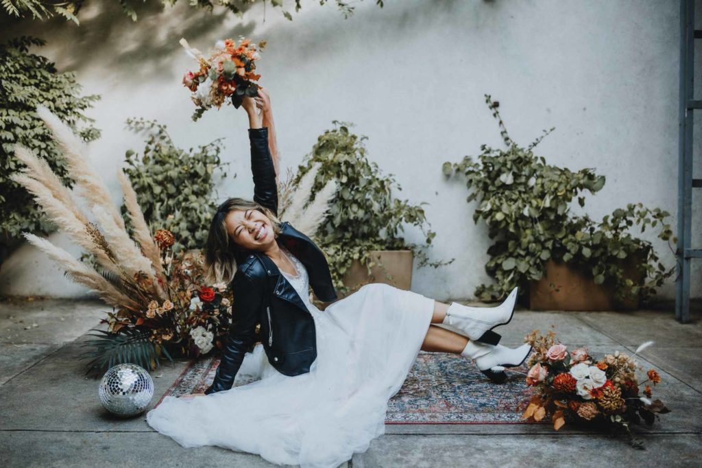 Outdoor Brooklyn microwedding styled shoot | Photography by Lucie B Photography | Featured on Equally Wed, the world's leading LGBTQ+ wedding magazine | marrier poses in BHLDN wedding gown and black leather jacket sitting by Boho decor and muted dusty rose florals