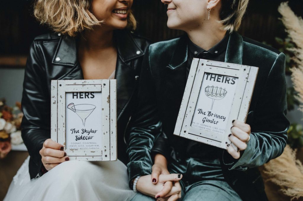 Outdoor Brooklyn microwedding styled shoot | Photography by Lucie B Photography | Featured on Equally Wed, the world's leading LGBTQ+ wedding magazine | LGBTQ+ couple holding their signature cocktail signs with their pronouns, HERS and THEIRS