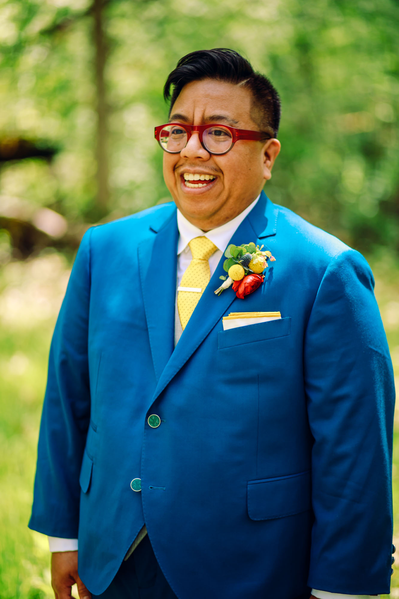 trans Brown man with red glasses and black hair in a blue suit, yellow tie and rainbow boutonniere