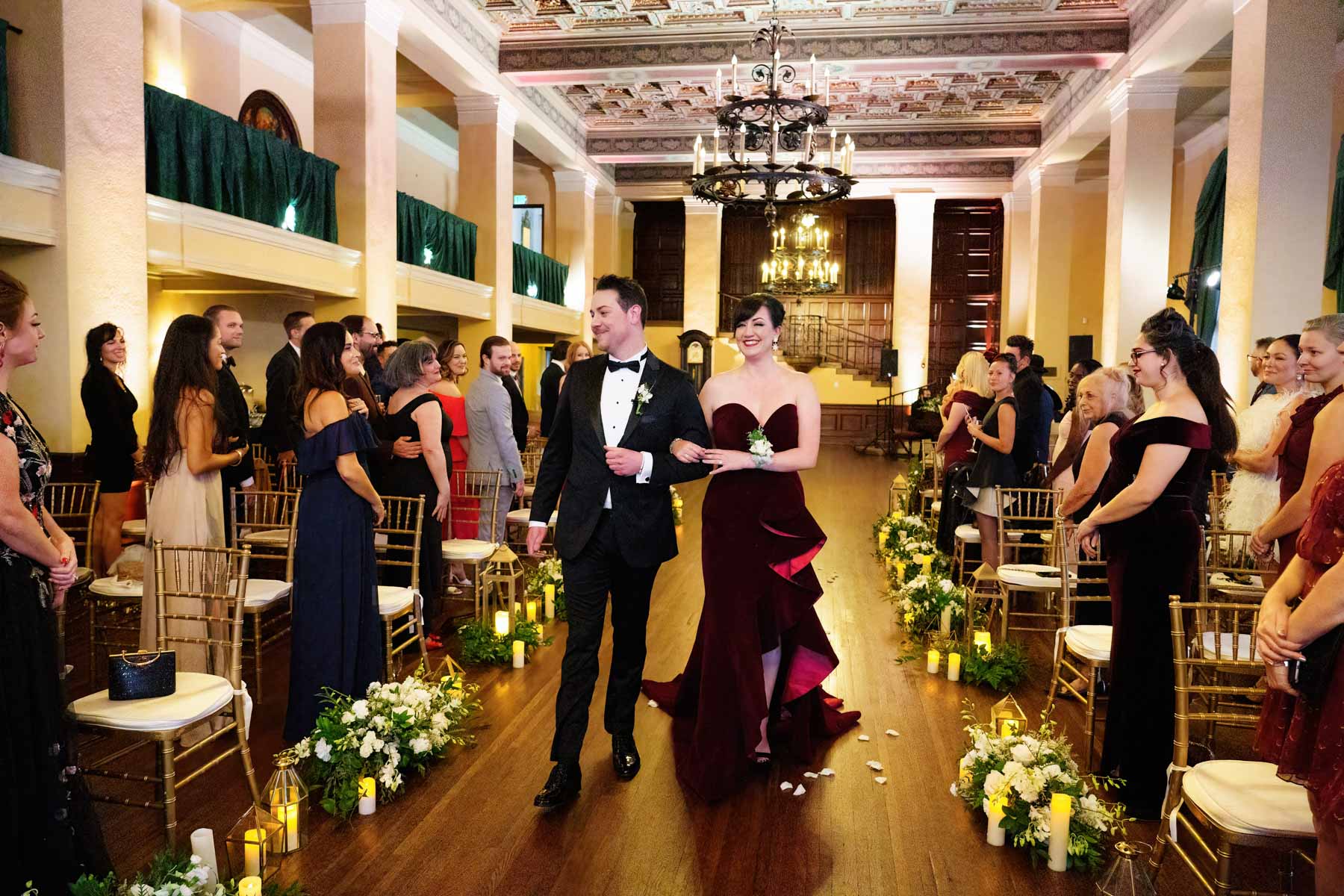 groom escorted up aisle with a woman in a strapless red dress while wedding guests watch
