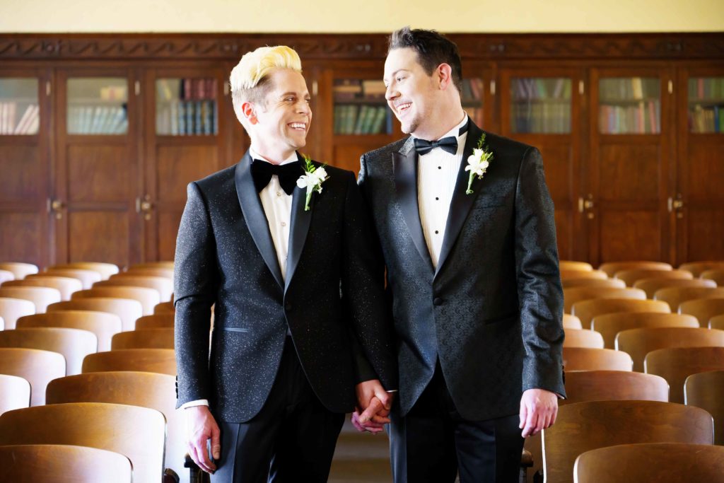 two grooms in black tuxedos smiling at each other