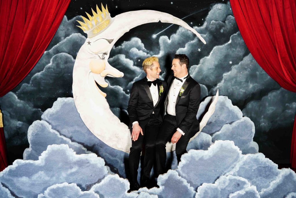 large moon installation for wedding photo booth with gay grooms sitting on it smiling at each other