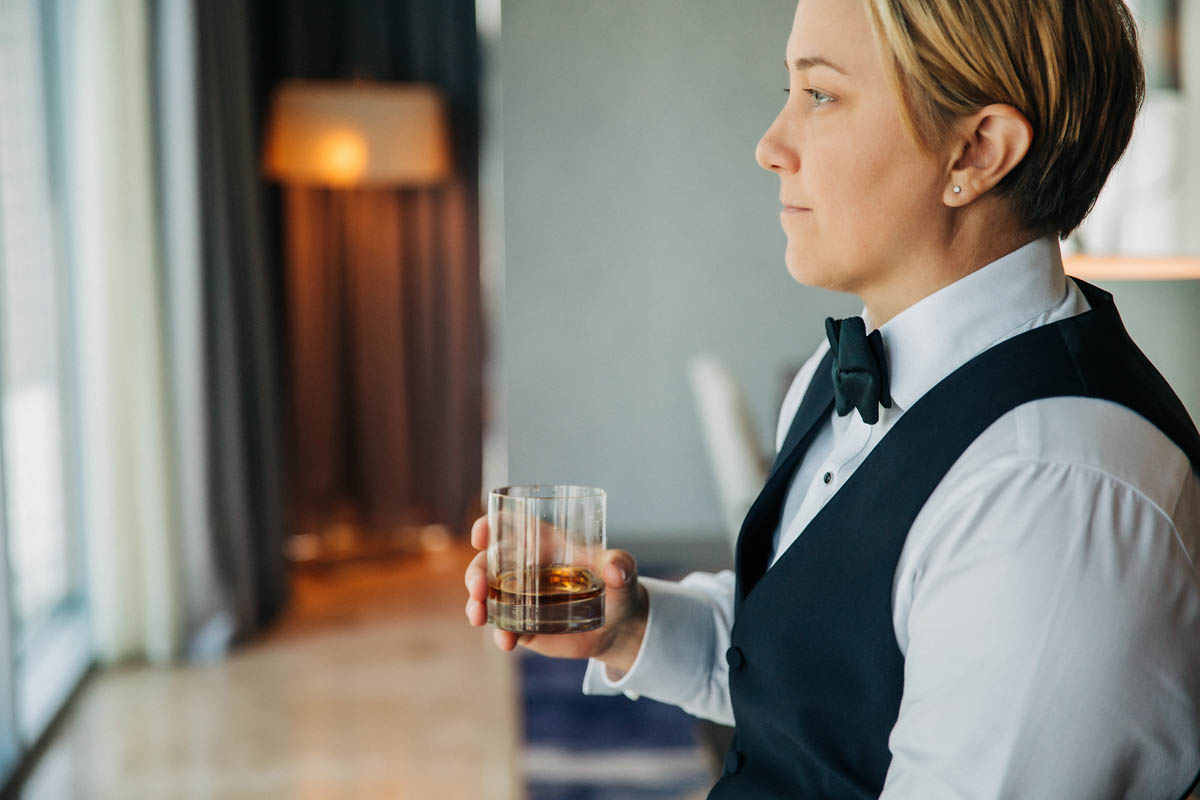 white nonbinary person in vest and bow tie with bourbon