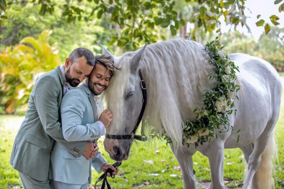 Two white grooms with brown hair and pale suits lean into each other while petting a white horse.
