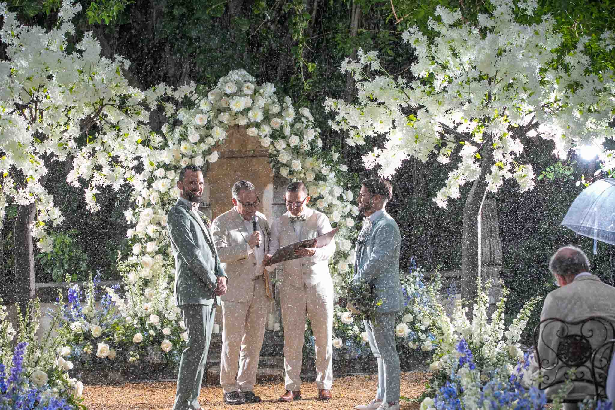 A pair of white grooms in light blue suits stand in the rain at their wedding ceremony, surrounded by white flowers and trees