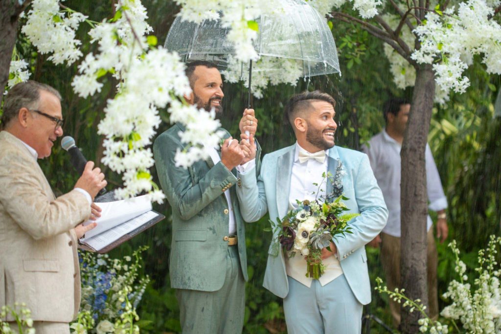 женихи на гей-свадьбе стоят под зонтиком, один жених держит букет. оба белые с каштановыми волосами.