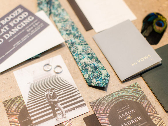 A flatlay of wedding day goods, including an invitation, tie, vow book, rings and ringboxes.