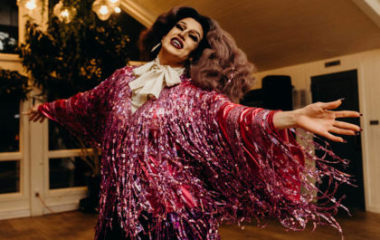 A drag queen in pink fringe dances in a reception hall.