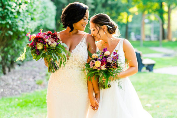 interracial couple holding hands and bouquets. one is a Black woman in a v-neck wedding gown and her wife, a white bride in a v-neck wedding gown is kissing her shoulder