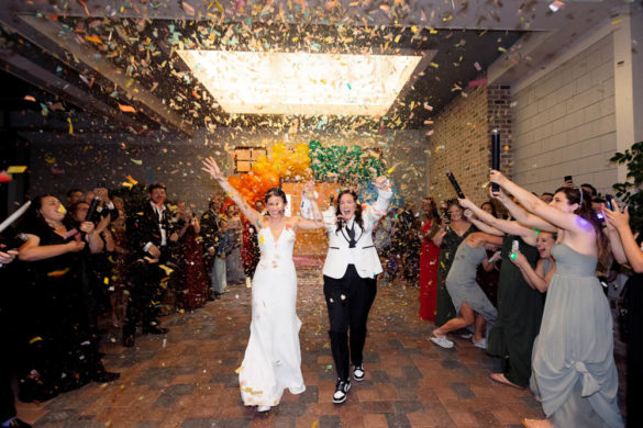 lesbian wedding of two white women walking through confettie being thrown by wedding guests. In the background is a rainbow balloon arch.