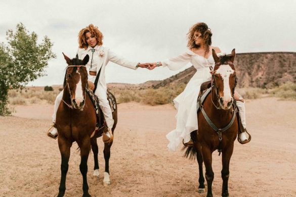 Lesbian brides sitting on brown horses in New Mexico desert. One marrier in a white suit with curly hair is looking at the camera and the other marrier is wearing an off-the-shoulder white wedding dress and is looking at her new wife.