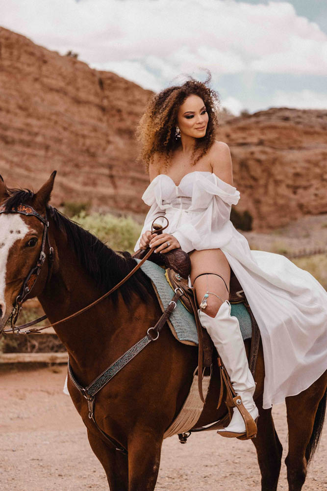 Lesbian bride sitting on horse. She's wearing an off-the-shoulder wedding dress and knee-high white leather boots.