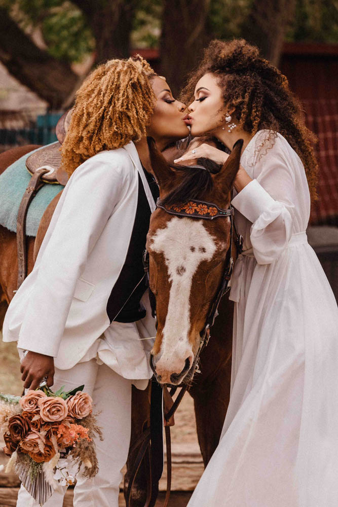 Lesbian couple kiss over a horse. Both have curly hair and are wearing white wedding attire.