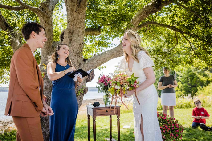 LGBTQ+ couple, one in a copper suit, one in an open-back lace wedding dress, say their vows. Brunette officiant in blue dress stands in the middle of them and is looking down at a notepad. Behind them is an apple tree and the ocean. LGBTQ+ couple, one in a copper suit, one in an open-back lace wedding dress, say their vows. Brunette officiant in blue dress stands in the middle of them and is looking down at a notepad. Behind them is an apple tree and the ocean.