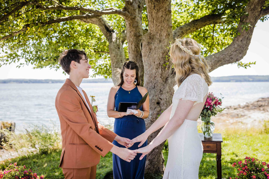 LGBTQ+ couple, one in a copper suit, one in an open-back lace wedding dress, say their vows. Brunette officiant in blue dress stands in the middle of them and is looking down at a notepad. Behind them is an apple tree and the ocean. LGBTQ+ couple, one in a copper suit, one in an open-back lace wedding dress, say their vows. Brunette officiant in blue dress stands in the middle of them and is looking down at a notepad. Behind them is an apple tree and the ocean.