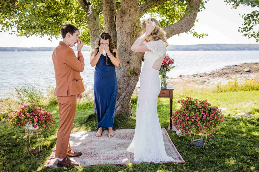 LGBTQ+ couple standing on a square rug at their wedding ceremony. Marrier with short curly brown hair and wearing a copper suit wipes their eyes. Brunette officiant in a blue dress and sandals dabs her eyes with a handkerchief. Blonde marrier in open-back ivory lace wedding dress adjusts her flower crown. Behind them is an apple tree and an ocean.