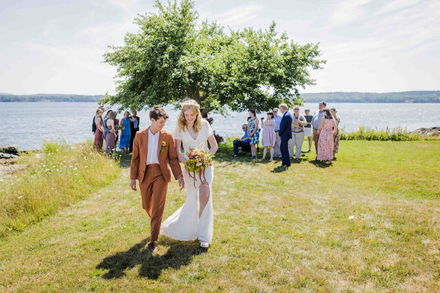 LGBTQ+ couple recessional at their outdoor summer wedding in Maine. Behind them is an apple tree and an ocean.