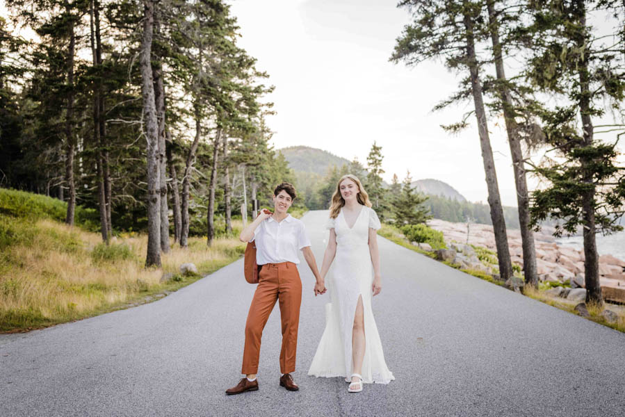 Smiling LGBTQ+ couple stands in middle of road holding hands. The brunette is wearing a white shirt and copper pants and the blonde is wearing a v-neck lace wedding dress with a slit and white sandals.