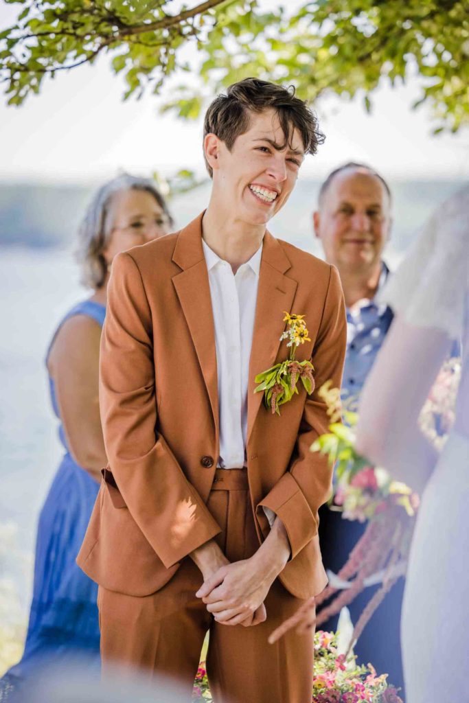 smiling genderfluid white person in copper suit with clasped hands listens as their fiancee says her vows at their outdoor summer wedding in Maine