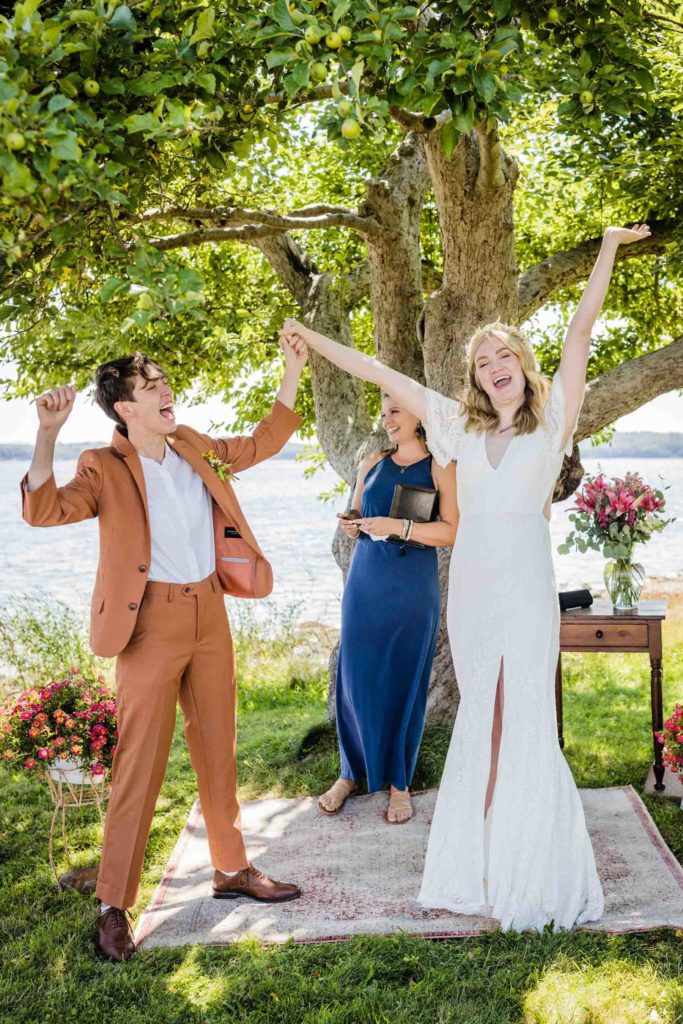 smiling white LGBTQ+ couple throw their hands in air after getting married under an apple tree at their outdoor summer wedding