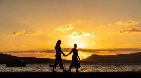 two women walk on beach at sunset in Greece with ocean in background, sky is yellow