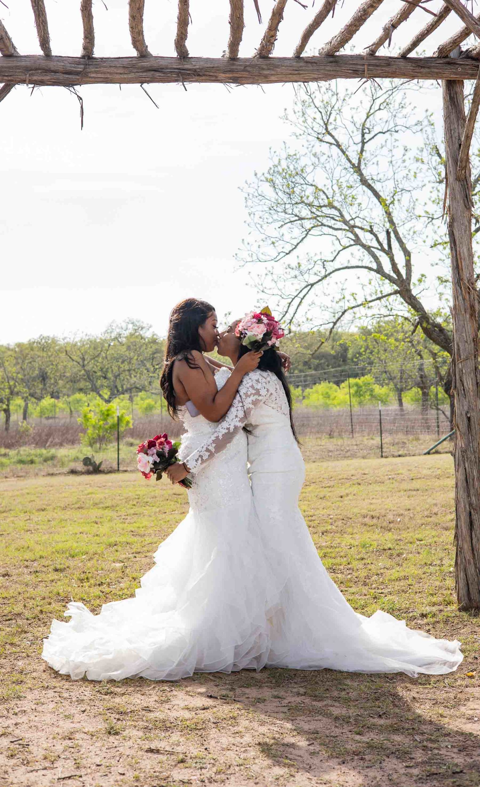 two Black lesbian brides kiss while wearing white wedding gowns and holding pink flower bouquets under wooden arch